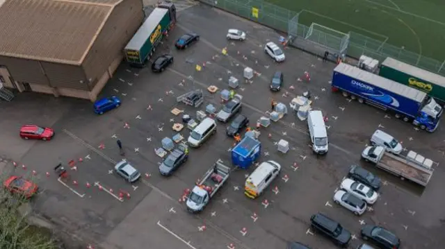 An aerial view of a water station as bottled water is loaded into cars at the emergency water distribution point in East Grinstead. Cars and trucks are lined up around cones and the white lines of parking spaces are visible in the car park.