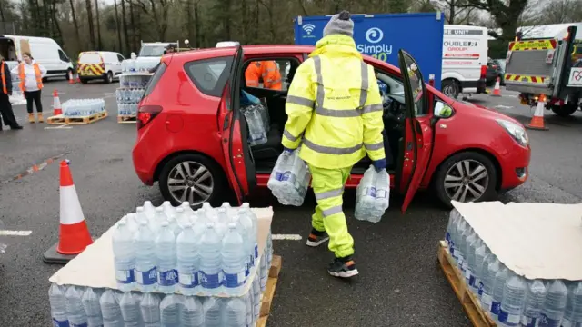 A person in a florescent jacket puts a dozen water bottles in a red car. More bottles are stacked up nearby