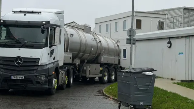 A water tanker stands near hospital buildings, parked near a grass verge.