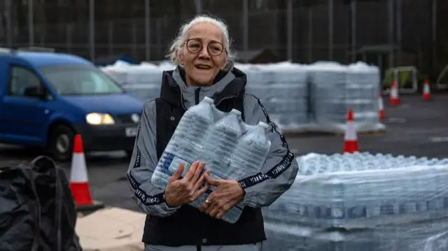 A woman collects bottled water from an emergency water distribution point in East Grinstead. She has grey hair tied back and is wearing glasses and an outdoor waterproof coat. She has a pack of six plastic bottles in her arms.