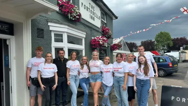 A family stand outside a pub. There's 11 of them, all wearing shirts that cheer on the England football team. There's English flags in the background.