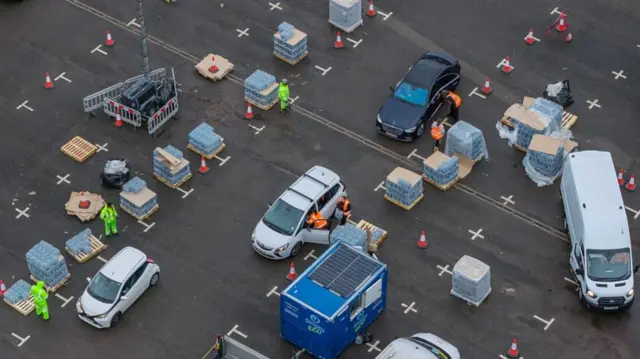 A distribution centre set up in a car park. There are crates full of bottled water set out in uniformed squares with cars passing to them through cordons.
