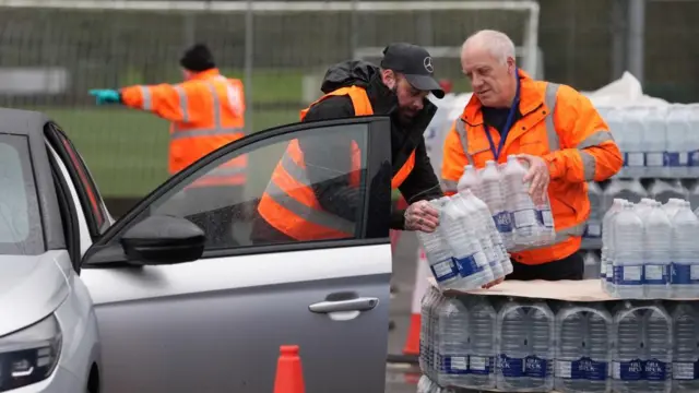 Two men in high vis jackets carry two six-packs of water to a car, whose door is open