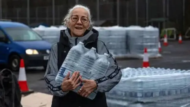 Elderly woman with white hair and glasses carrying a pack of six bottles of plastic water outdoors