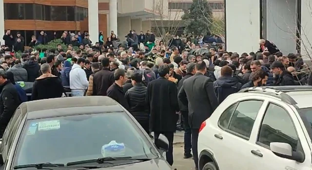 Hundreds of people waiting around in what seems like a car park outside a building. There are two cars parked in the foreground.