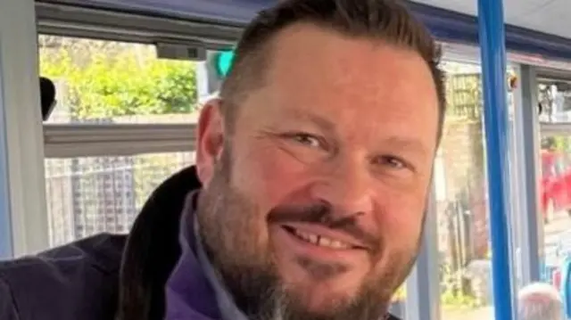 A man on a bus smiles to the camera. He's wearing a purple fleece and a coat.