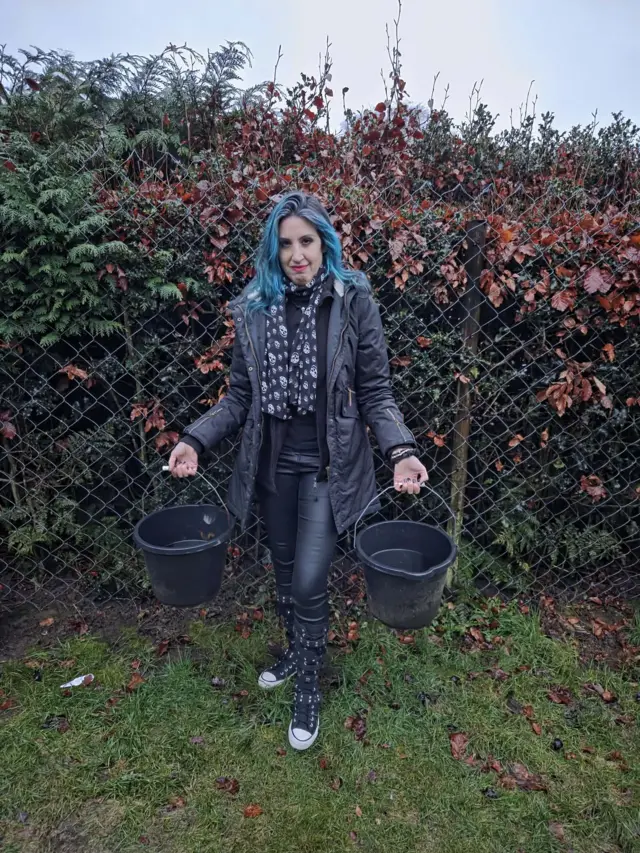 A woman with blue hair holds two buckets of water in her garden. She's wearing a black coat and jeans
