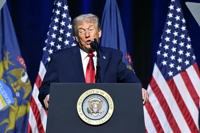 US President Donald Trump speaking behind a podium in a blue suit with red tie.