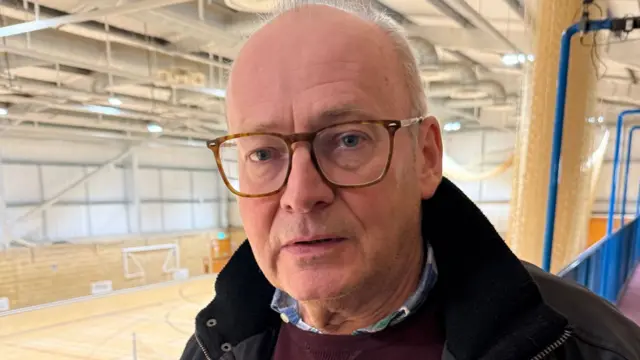 A bald man with glasses stood in a viewing platform overlooking an empty sports hall. He is looking into the camera and is wearing a black jacket