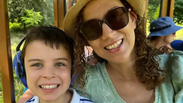 A boy wearing ear defenders is smiling next to his mum, who is wearing sunglasses and a straw hat. They're hugging and look really happy