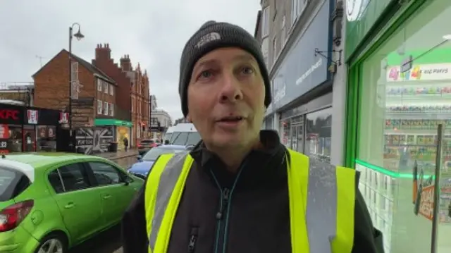 A man in a high-vis vest and black jumper wearing a black hat with a white logo on it. He is stood outside shops in a street, looking just above the camera