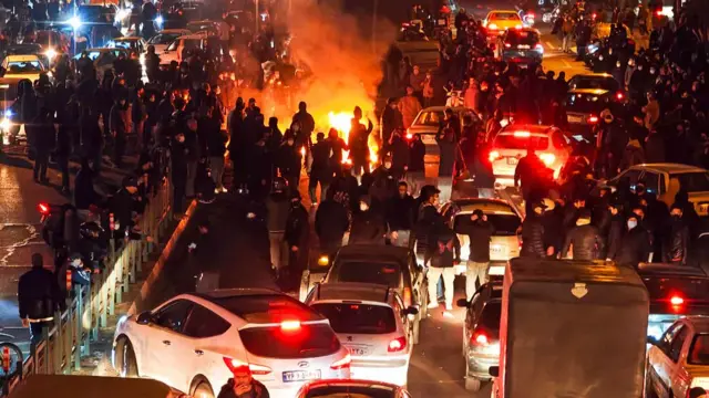 Protesters staanding on road at night, among the cars. something has been set alight on the road.