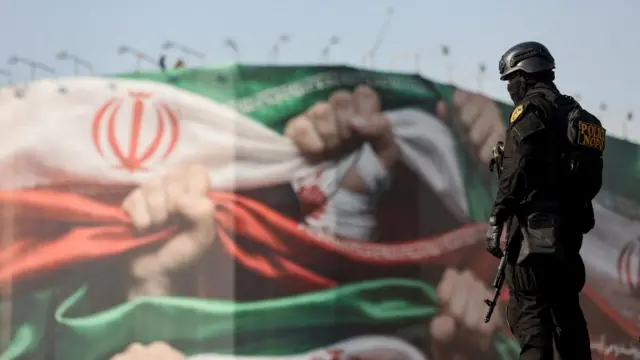 A police officer stands in front of a painted wall which shows hands clutching the Iranian flag