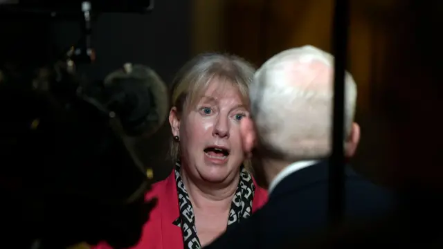 Shona Robison addresses a journalist with white hair who is facing away from the camera and towards her. He stands next to a camera pointed at Robison. She has tied back fair hair and wears a pink blazer and black and white shirt.