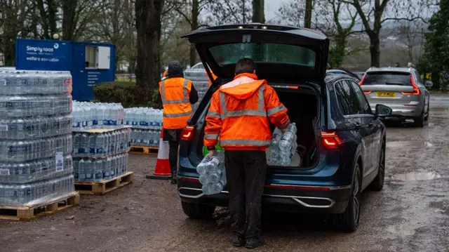 Two people in high vis fill a car boot with bottles of water. We can see a queue of cars also being served. There are lots of crates full of water bottles in the background.