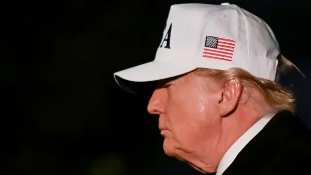 A side view of Donald Trump wearing a white cap with a USA flag on it.