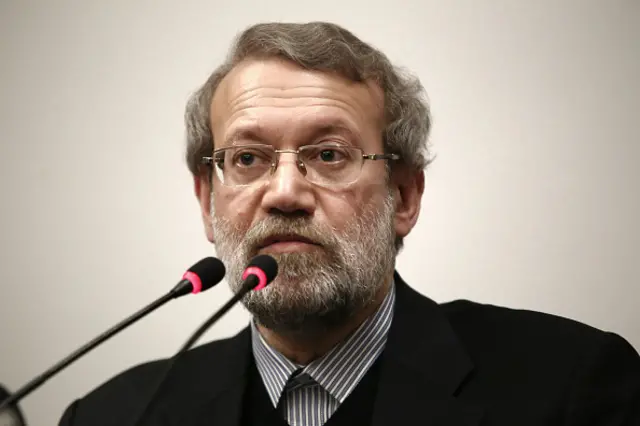 Ali Larijani speaking into two black and pink microphones, wearing glasses, a buttoned-up stripey grey and white shirt and a black blazer