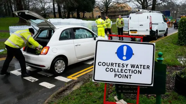 A car and a van stopped on a road next to pallets of bottled water. The white car has its boot open and a person in a high-vis jacket is reaching into the boot.