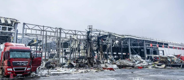 A heavily damaged postal depot building and lorry, there is burn marks, rubble strewn around and part of the building has lost much of its structure with just metal frames left standing