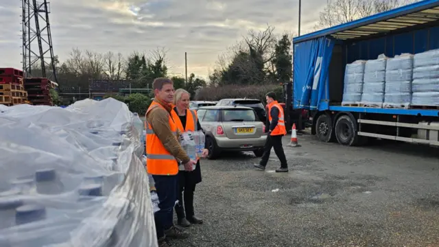 Two people carrying cases of water and stood beside a large pallet of bottles. Cars can be seen behind them, as can multiple pallets in a curtained lorry trailer.
