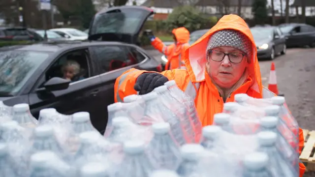 A woman in an orange jacket handles bottles of water.