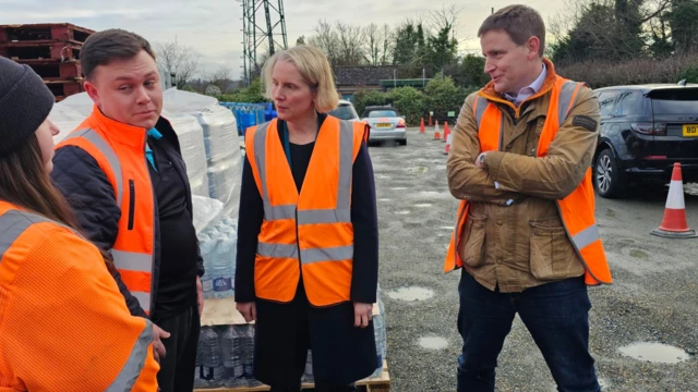 Four people talking while wearing orange high-vis jackets. Cars and large pallets of water can be seen behind them.