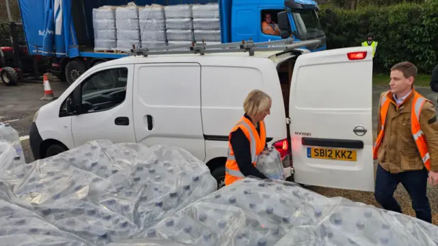 A woman carries a case of bottled water beside pallets of bottles towards the open door on the back of a small, white van. A man is stood at the back of the van. Both people are wearing high-vis jackets.