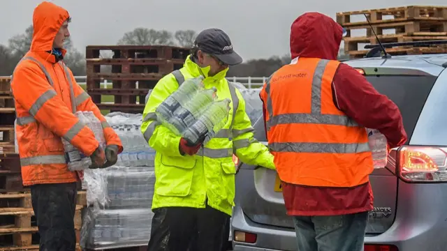 Three people in high-vis carrying cases of water at the back of a car. Stacks of empty pallets are seen in the background.