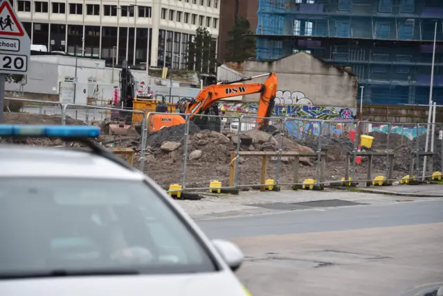 A building site inside a cordon in Plymouth. There is a police car in the foreground and a muddy building site with an orange digger in the background.