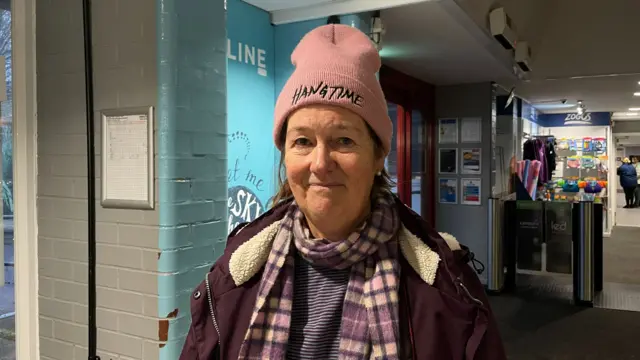 A woman wearing a hat, scarf and coat smiles at the camera. She is standing inside a leisure centre building. Swimming goggles and woggles are on display behind her.