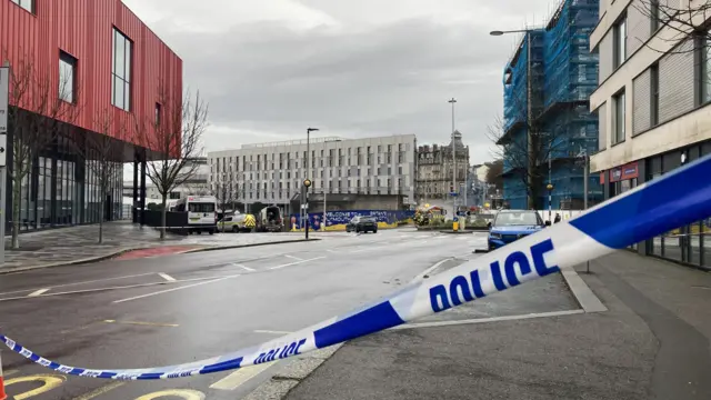A road closed off by police tape, showing police vehicles and vans in the distance.