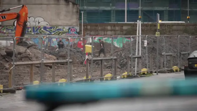 A building site in Plymouth with an orange digger and workers behind a metal fence