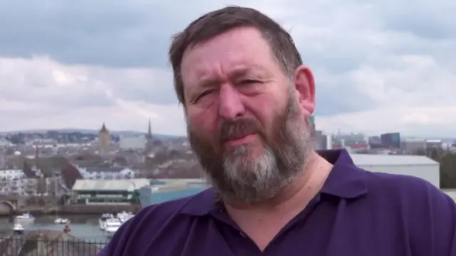 Dr Harry Bennett, looking towards the camera wearing a purple polo shirt which says 'HMS Gloucester' on it