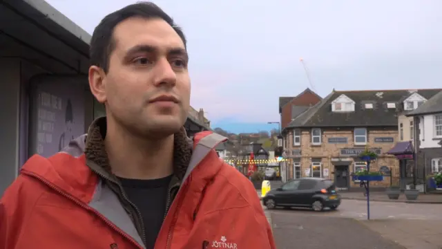 A man in a red coat and black t-shirt. He is looking to the right but facing forward. Behind him is a car driving past. The sky is blue with white clouds.