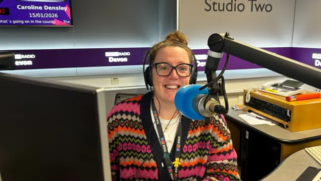 Caroline Densley inside the BBC Radio studio. She is wearing a cardigan and headphones and glasses. She has a microphone in front of her.