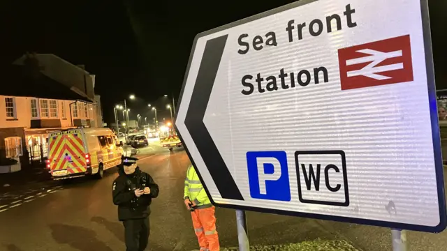 A sign for the sea front pointing to the road. There is a police van and highways van on the road. There is a police officer and a person stood in high-vis clothing near the sign.
