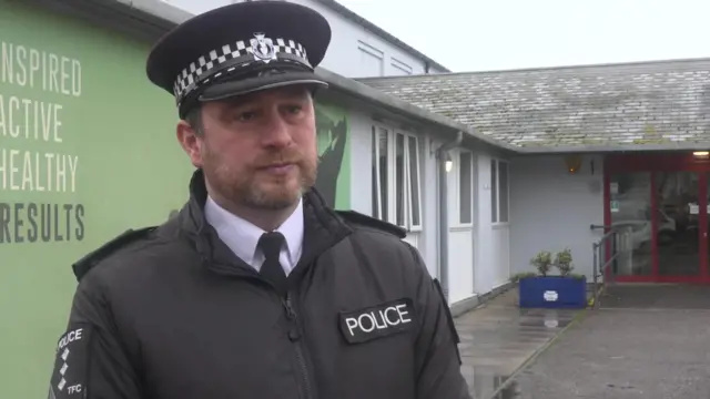 A man in police uniform. He is looking towards the camera. Behind him is a building. The sky is grey.