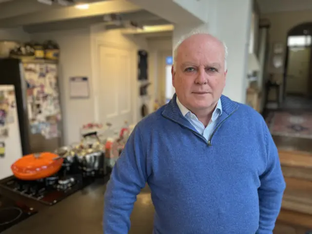 A man wearing a blue jumper and shirt smiles at a camera. He is standing indoors. A kitchen set up is behind him.