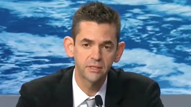Jared Isaacman in a black suit, white shirt and grey tie, sitting at a desk in front of a microphone.