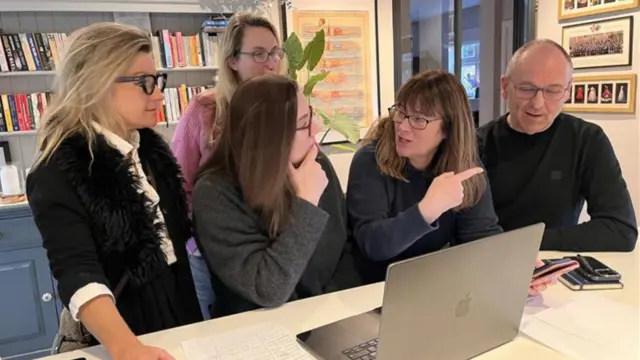 Five people in conversation around an open laptop. They are in a room with bookshelves and framed art on the walls.