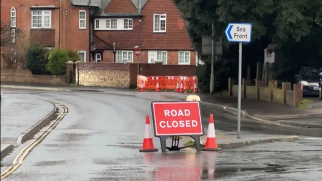 A road closure sign on display in the middle of a road. A sign next to it points to the seafront. Properties line the road.