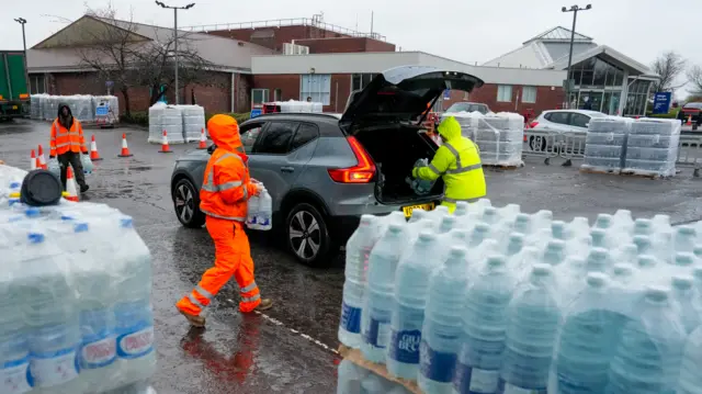 People in high vis jackets loading bottled water into a car with its boot open.
