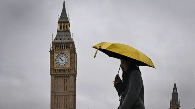Elizabeth Tower at the Houses of Parliament in London. The sky is dark and a woman with a yellow umbrella walks past the building.