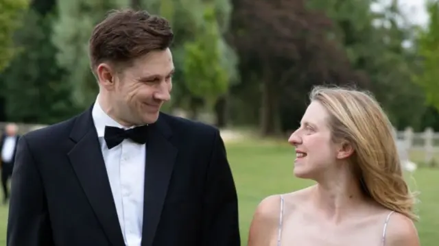 A man in a black tuxedo and a woman with long, blonde hair smiling and looking at eachother.