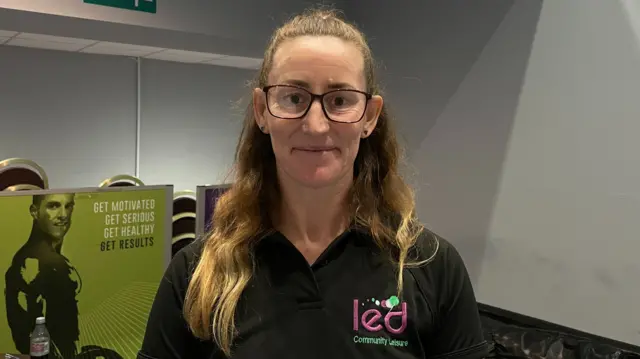 Emma Kessie in her black staff uniform in the leisure centre. She has long tied back hair and glasses