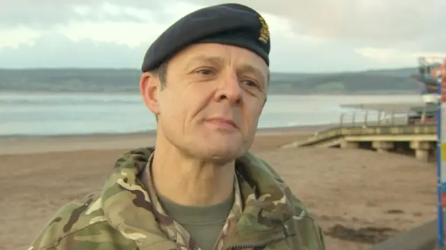 A man in military uniform looking towards the camera. He is stood in front of a beach.