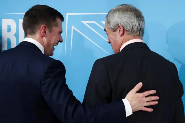Jenrick and Farage, shown from behind. Jenrick has his arm on Farage's back and they are both laughing and looking at eachother.