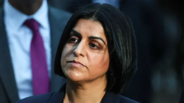 Shabana Mahmood looking into distance and wearing a dark blue suit with a gold necklace, with an unrecognisable man in a suit behind her