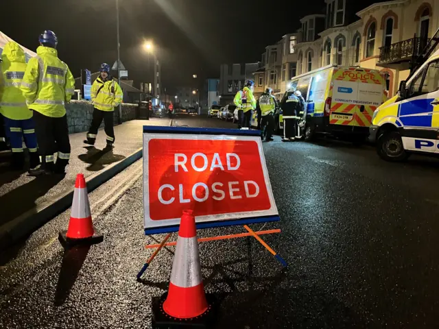Various emergency services and workers behind cordon tape and a road closed sign