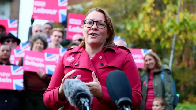 Turley speaking into a mic at a Labour event, with a crowd behind her holding posters reading 'RENEW BRITAIN'
